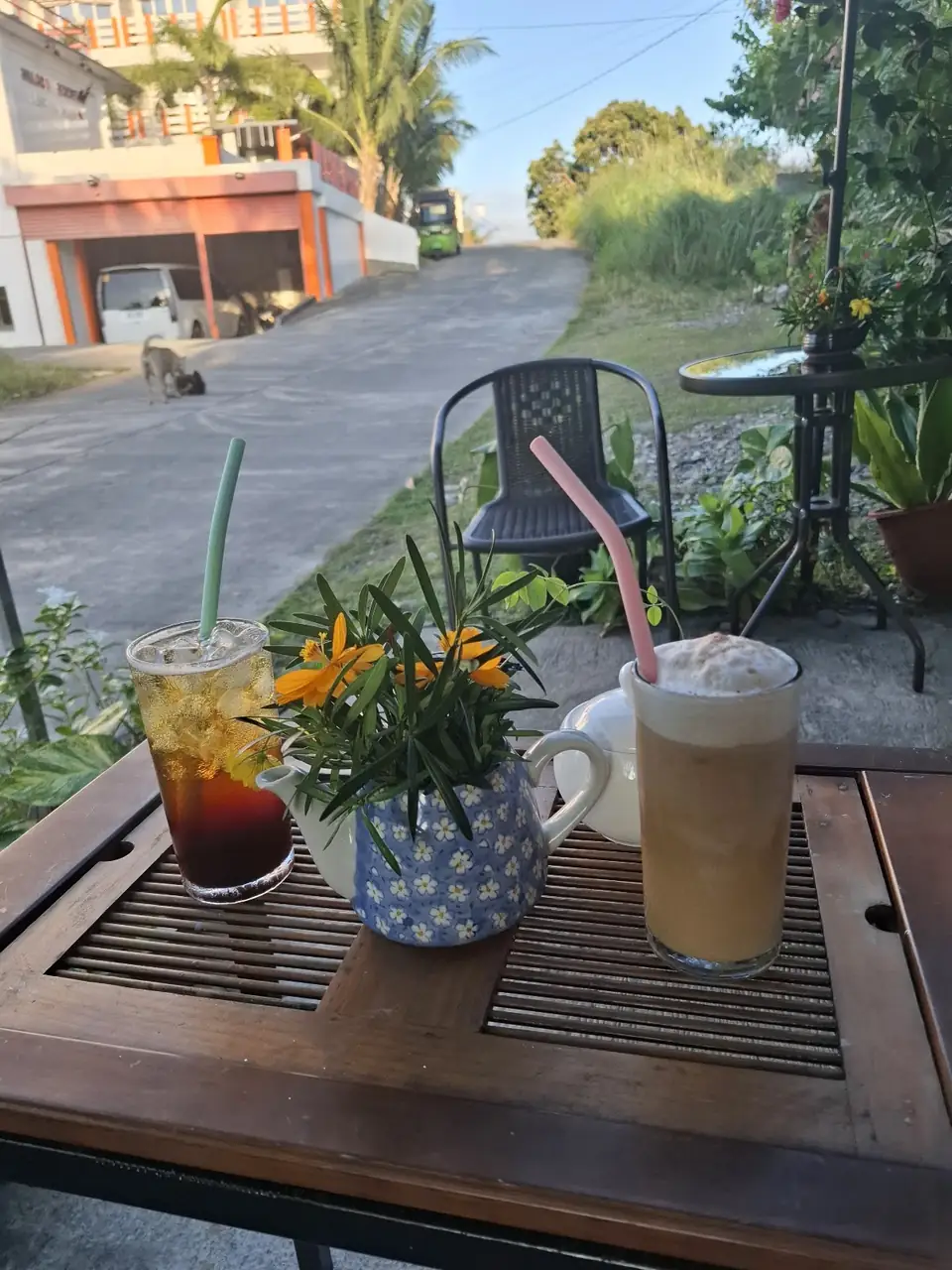 An iced dark drink and a tall foamy coffee on a wooden table with orange flowers